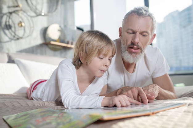 Nauczanie, czytanie. Poważny siwowłosy tata uczy dziecko czytać, leżąc na łóżku w domu, przesuwając palcami po książce
