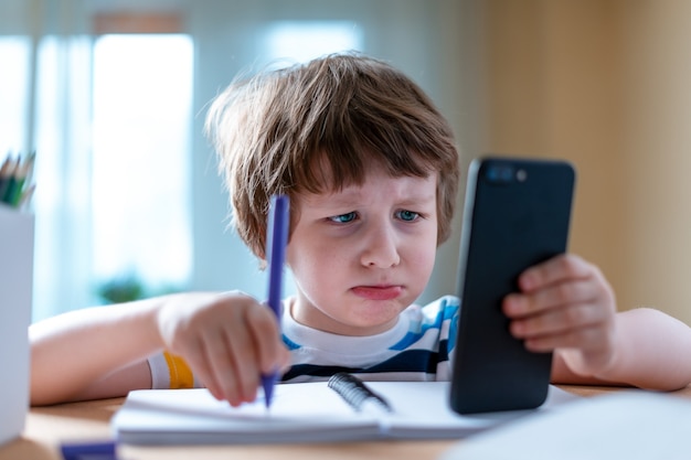 Nauczanie na odległość edukacja online kaukaski smutny chłopiec uczy się w domu ze smartfonem i robi