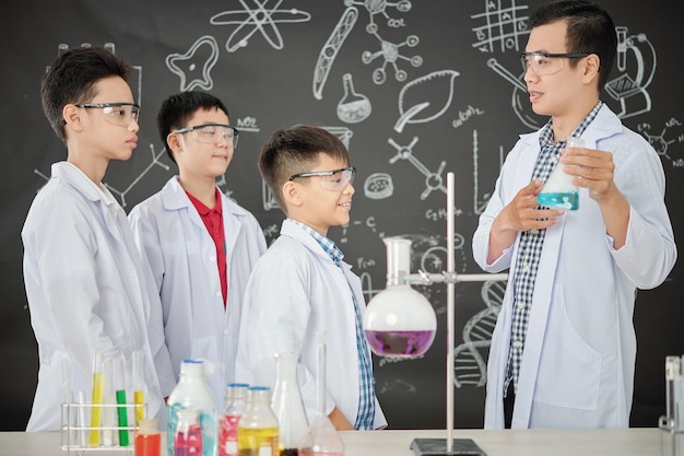 Nauczyciel chemii pokazuje grupie podekscytowanych uczniów w okularach i fartuchach zlewkę z niebieskim płynem