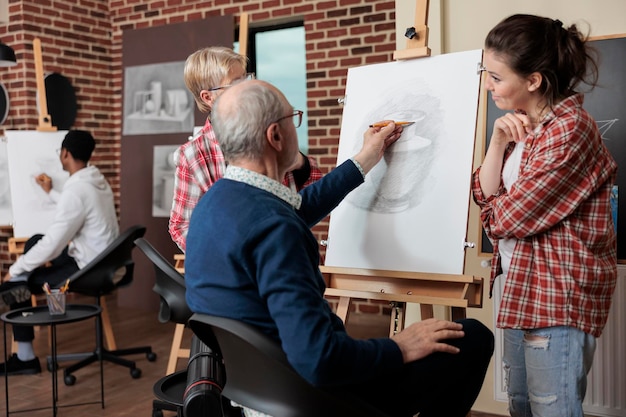 Nauczyciel nadzorujący starszego ucznia podczas rysowania modelu wazonu na płótnie pracujący w technice ilustracji w studio kreatywności. Zróżnicowany zespół uczęszczający na zajęcia plastyczne rozwijające umiejętności artystyczne