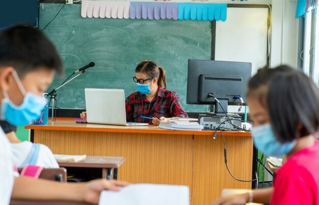 Nauczyciel Noszący Maskę Ochronną, Aby Chronić Się Przed Covid-19, Uczy Dzieci W Szkole Siedzących W Klasie Online, Szkoła Podstawowa, Nauka I Koncepcja Ludzi.