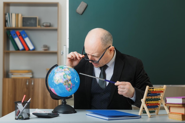 Nauczyciel w okularach siedzi z kulą ziemską przy ławce szkolnej przed tablicą w klasie, wyjaśniając lekcję z zaintrygowaną miną