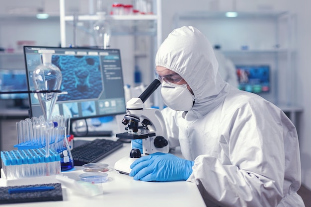 Naukowiec chemii patrząc na próbkę na szkiełku za pomocą mikroskopu. Virolog w kombinezonie podczas epidemii koronawirusa przeprowadzający naukowe analizy opieki zdrowotnej.