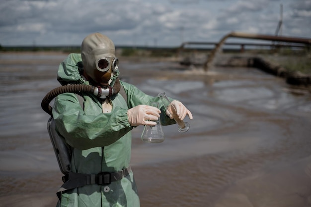 Zdjęcie naukowiec pobiera próbkę wody ze zbiornika po uwolnieniu odpadów chemicznych mężczyzna w respiratorze i zielonym kombinezonie ochronnym przed promieniowaniem eksperci analizy wody maska