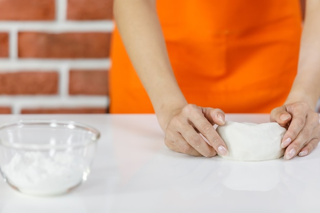 Nie do poznania kobieta w pomarańczowym fartuchu uśmiecha się i pochyla, aby cieszyć się wyrabianiem ciasta rękami obok szklanej miski mąki w proszku na białym stole do gotowania jako przygotowanie jedzenia w pobliżu ceglanego muru w domowej kuchni