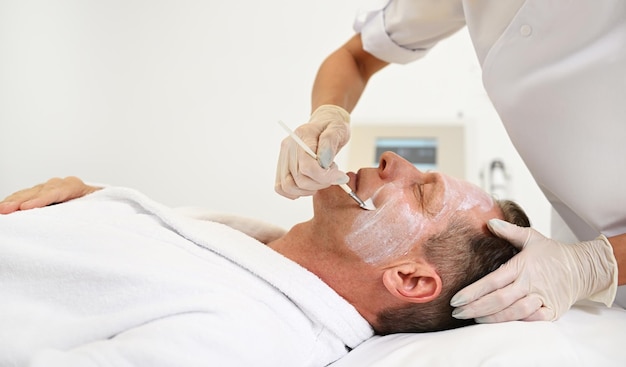 Nie do poznania kosmetyczka stosująca kosmetyk kosmetyczny pędzlem na twarzy przystojnego dojrzałego mężczyzny leżącego na stole do masażu podczas odbierania męskiej profesjonalnej pielęgnacji skóry w spa wellness