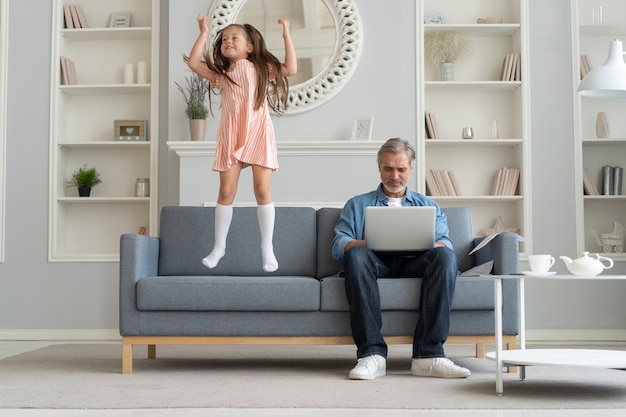 Nie ma czasu dla dziecka. Siwowłosy tata zajęty laptopem, pracujący online w domu, smutna znudzona obrażona córka skacząca w pobliżu.