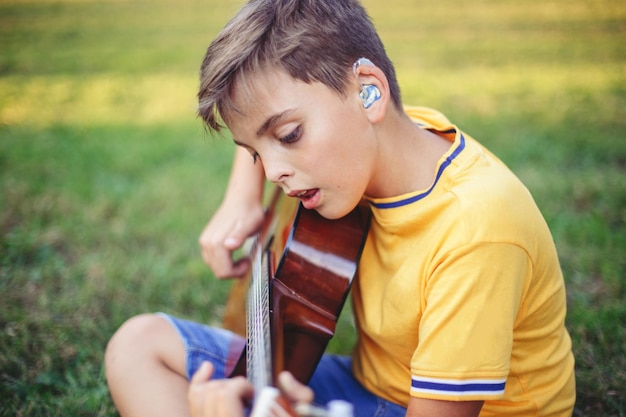 Zdjęcie nie słyszący nastoletni chłopiec grający na gitarze i śpiewający dziecko z aparatami słuchowymi grające muzykę