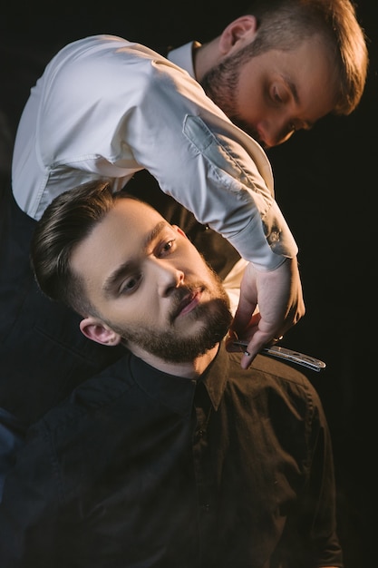 Niebezpieczne męskie golenie ostrą brzytwą. Zbliżenie w stylu współczesnego mężczyzny, fryzjer z czarnym tłem, uważny stylowy fryzjer, męska koncepcja