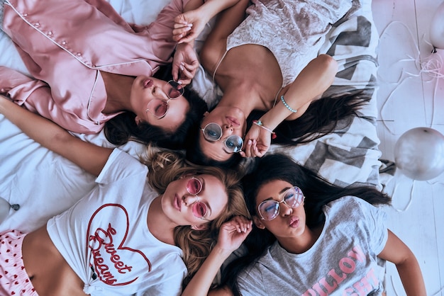 Niebezpiecznie piękna. Widok z góry na cztery zabawne młode kobiety w okularach, uśmiechnięte i robiące minę, leżąc na łóżku w domu