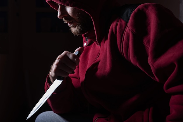 Niebezpieczny brodaty mężczyzna w czerwonym kapturze trzyma ostry nóż blisko twarzy Przestępca lub zabójca z nożami w ciemności