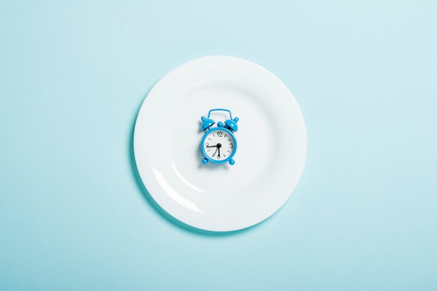 Niebieski budzik leży na białym talerzu na niebieskiej ścianie. Pojęcie diety, harmonogram posiłków, utrata masy ciała. Leżał płasko, widok z góry