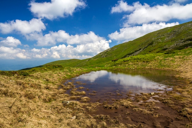 Niebieskie Niebo Z Jaskrawymi Białymi Chmurami Odbijał W Małym Jeziorze Między Zielonymi Wzgórzami Na Słonecznym Dniu. Letnia Panorama.