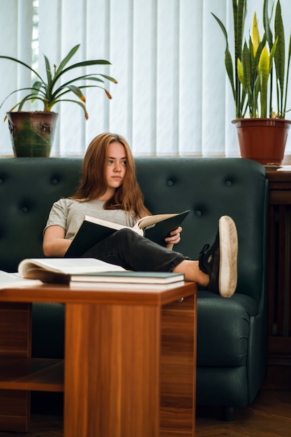 Niebieskooka Ruda Dziewczyna Z Poważnym Wyrazem Twarzy Odpoczywa W Bibliotece Publicznej Na Kanapie Z Otwartą Książką Na Kolanach I Nogą Na Stole, Odwracając Wzrok.