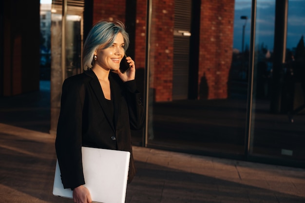 Niebieskowłosy kaukaski przedsiębiorca rozmawiający przez telefon podczas spaceru z komputerem