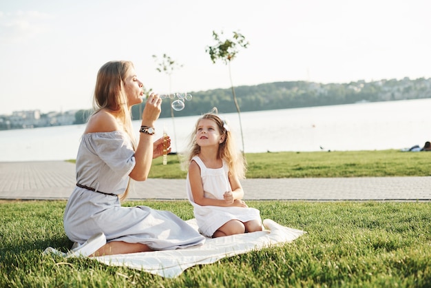 Niech nadejdzie wystrzelenie bąbelków. Zdjęcie młodej matki i jej córki, dobrą zabawę na zielonej trawie z jeziorem w tle.