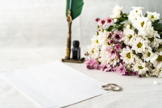 Niektóre pierścionki ślubne lub zaręczynowe wraz ze starym piórem w kałamarzu i białą kartką gotową do napisania notatki. Tej kompozycji towarzyszą piękne kwiaty.