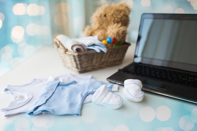 niemowlęctwo, macierzyństwo, odzież, technologia i koncepcja obiektu - zbliżenie ubrań dla niemowląt i zabawek dla nowonarodzonego chłopca w koszu z laptopem w domu