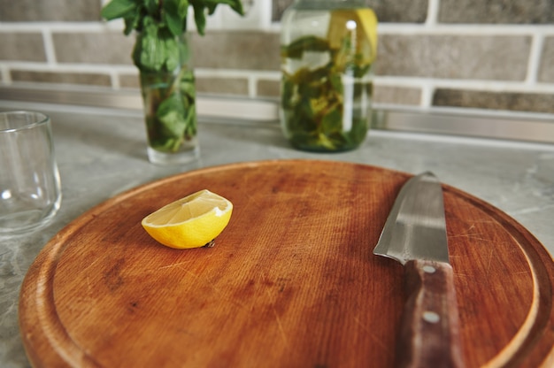 Nieostrość na połówce cytryny nożem na drewnianej desce na blacie kuchennym na tle zamazanej butelki z wodą cytrynowo-miętową i szklanką z liśćmi mięty