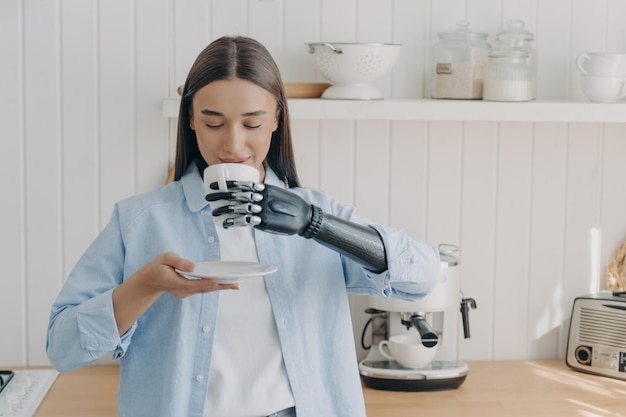Niepełnosprawna dziewczyna pijąca kawę lub herbatę trzymającą kubek za pomocą zaawansowanej technologicznie bionicznej protezy ręki w kuchni