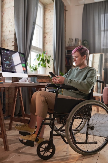 Niepełnosprawny grafik siedzący na wózku inwalidzkim przed swoim miejscem pracy z komputerem i rozmawiający przez Internet przez telefon