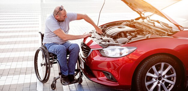 Niepełnosprawny mężczyzna na wózku inwalidzkim, sprawdza silnik swojego samochodu na parkingu