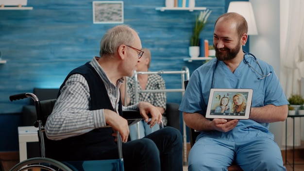Zdjęcie niepełnosprawny mężczyzna rozmawia wideorozmowę z rodziną w domu opieki. asystent medyczny z tabletem pomagający staremu pacjentowi w zdalnej komunikacji podczas konferencji online z krewnymi