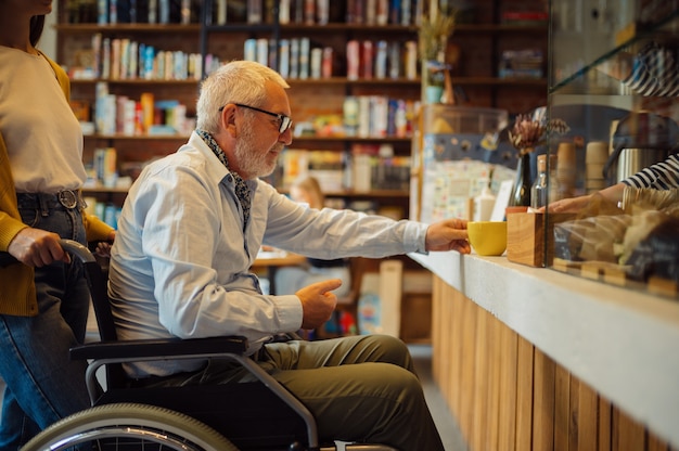 Niepełnosprawnych dziadek na wózku inwalidzkim i wnuczka w kasie, niepełnosprawność, wnętrze kawiarni na tle. Niepełnosprawny starszy mężczyzna i młoda opiekunka, sparaliżowani ludzie w miejscach publicznych