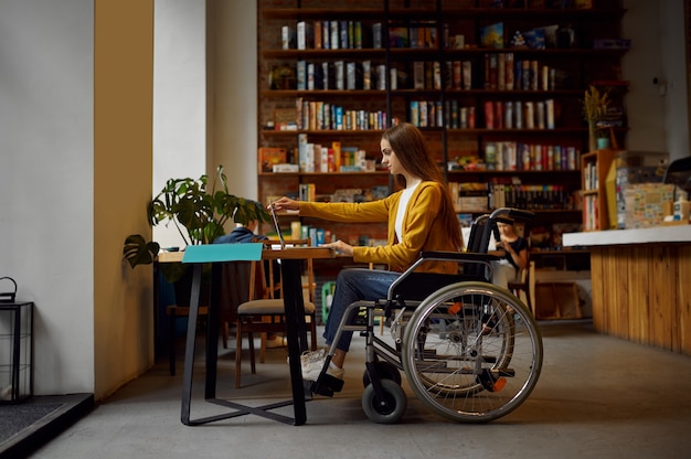 Niepełnosprawnych studentka na wózku inwalidzkim za pomocą laptopa, niepełnosprawności, regału i wnętrza biblioteki uniwersyteckiej na tle. Niepełnosprawna młoda kobieta studiująca na studiach, sparaliżowani ludzie zdobywają wiedzę