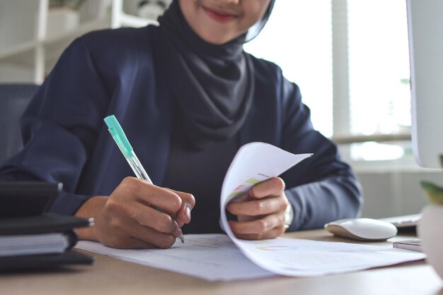 Nierozpoznawalna azjatycka muzułmanka w chustce na głowie pracująca przy stole biurowym, pisząca coś w niej