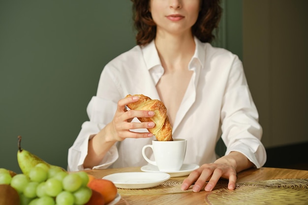 Nierozpoznawalna kobieta zanurzająca rogalika w cappuccino rano w domu