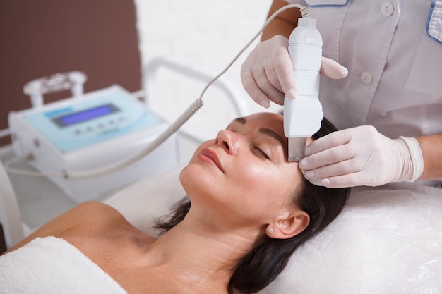 Nierozpoznawalny kosmetolog wykonujący ultradźwiękową pielęgnację twarzy dla dojrzałej kobiety