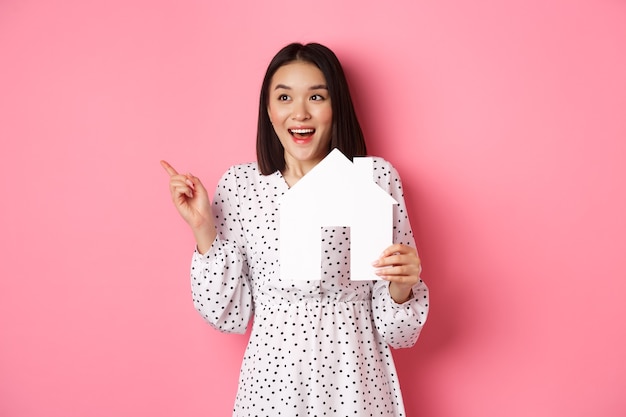 Nieruchomość. Podekscytowana azjatycka kobieta pokazująca papierowy model domu, wskazując i patrząc w lewo na miejsce, stojąc na różowym tle
