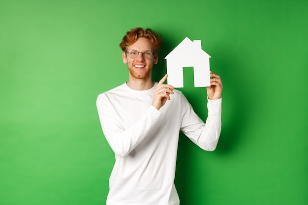 Nieruchomość. Przystojny młody mężczyzna z rudymi włosami, w okularach, pokazując wycinankę domu papieru i uśmiechnięty, stojąc na zielonym tle.