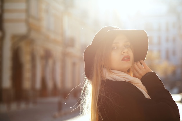 Niesamowita blondynka w kapeluszu, szaliku i płaszczu, idąc starą ulicą w promieniach słońca. Miejsce na tekst