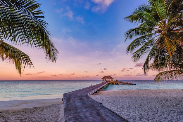 Niesamowita panorama zachodu słońca na Malediwach. Luksusowe wille wypoczynkowe nad brzegiem morza z liśćmi palmowymi i molo