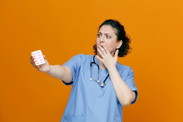 Niespokojna lekarka w średnim wieku nosząca mundur i stetoskop na szyi, rozciągająca paczkę kapsułek, patrząc na nią, trzymając rękę na ustach na białym tle na pomarańczowym tle