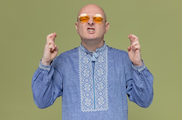 Niespokojny dorosły słowiański mężczyzna w niebieskiej koszuli w okularach przeciwsłonecznych krzyżujących palce