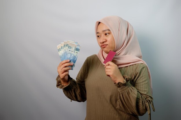 Nieszczęśliwa piękna azjatycka muzułmańska kobieta trzymająca papierowe pieniądze i pokazująca kartę kredytową odizolowaną na białym tle