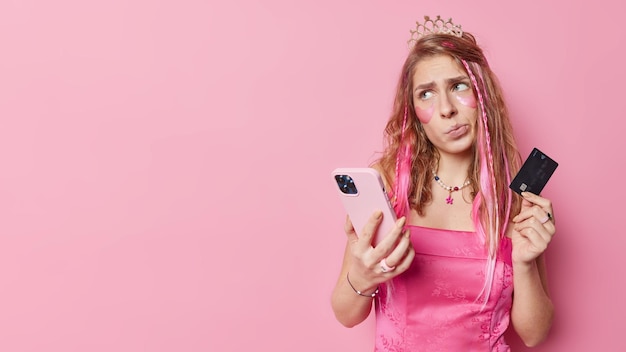 Nieszczęśliwa Zamyślona Młoda Kobieta Wygląda Na Niezadowoloną Odwraca Wzrok Trzyma Telefon Komórkowy I Kartę Kredytową Sprawia, że Zakupy Online W świątecznych Ubraniach Izolowanych Na Różowym Tle Puste Miejsce Na Promo