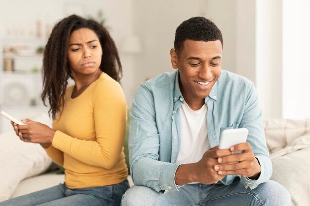 Nieszczęśliwa zazdrosna młoda Afroamerykanka ze smartfonem patrzy na męski telefon we wnętrzu pokoju