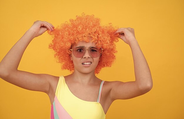 Nieszczęśliwe dziecko w okularach przeciwsłonecznych i kostiumie kąpielowym nie lubi pomarańczowej peruki na żółtym tle nie lubi