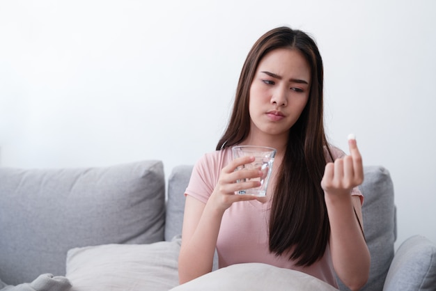 Nieszczęśliwy chory azjatykci młodej kobiety mienia lek i szkło wodny środek przeciwbólowy uśmierzać ból brzucha podczas gdy siedzący na kanapy kanapie w żywym pokoju w domu.