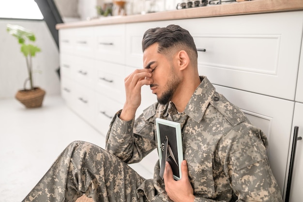 Nieszczęśliwy wojskowy siedzący na podłodze w kuchni i przyciskający zdjęcie do piersi