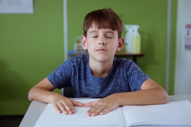 Niewidomy kaukaski uczeń siedzi w klasie z zamkniętymi oczami czytając książkę Braille'a palcami