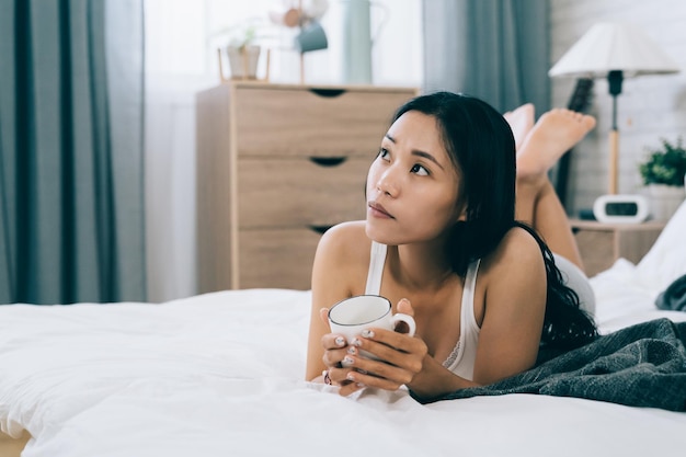 niewyraźne tło całkiem azjatyckie dziewczyny leżącej na łóżku z kawą jest znudzony. portret młodej kobiety koreańskiej na luźnym końcu w jej sypialni.