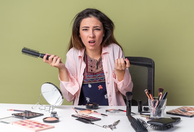 Niezadowolona młoda brunetka siedzi przy stole z narzędziami do makijażu, trzymając grzebienie izolowane na oliwkowozielonej ścianie z miejscem na kopię