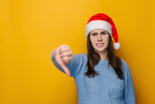Niezadowolona młoda kobieta pokazuje znak dezaprobaty, trzyma kciuk w dół, wyraża niechęć, marszczy czoło w niezadowolonej, ubrana w świąteczną czapkę, odizolowana na żółtym tle. Koncepcja wakacje nowy rok