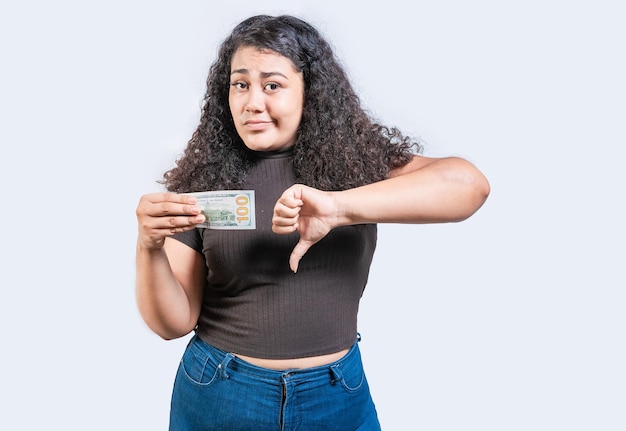 Niezadowolona młoda kobieta trzymająca banknot dolarowy z kciukiem w dół odizolowana smutna dziewczyna trzymująca banknot 100 dolarów z kciuki w dół