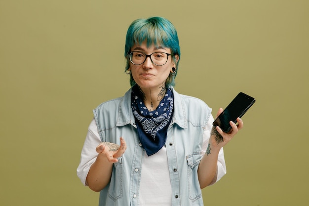 Niezadowolona Młoda Kobieta W Okularach Bandana Na Szyi, Trzymająca Telefon Komórkowy, Patrząca Na Kamerę Pokazującą Pustą Rękę Odizolowaną Na Oliwkowozielonym Tle